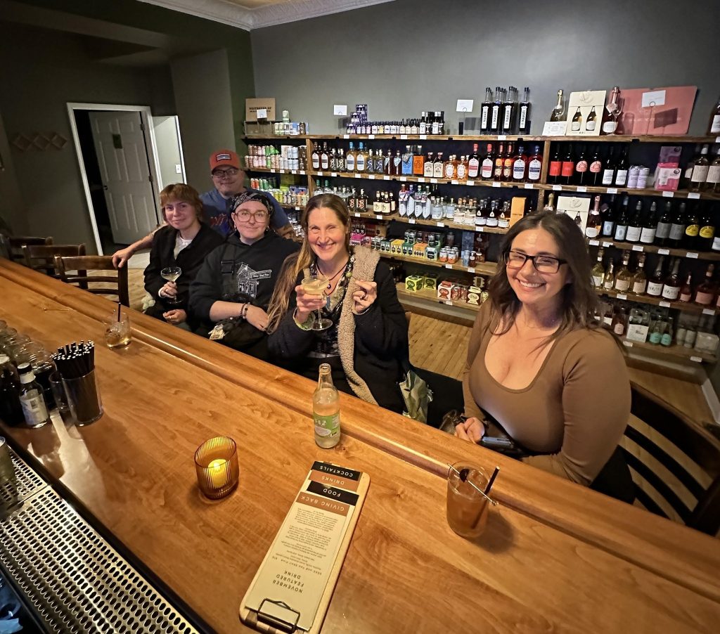 The image shows a group of five people sitting at a wooden bar counter at Alt-Bar. Behind them, there is a wall filled with shelves stocked with various bottles of non alcoholic bottled wine. The people are smiling and appear to be enjoying themselves, with some holding drinks. The person closest to the camera is a woman with glasses and long hair, wearing a brown top. The atmosphere seems relaxed and social.